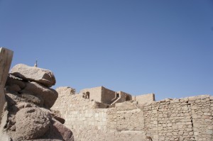 The image shows ancient stone ruins under a clear blue sky. The structures are made of large, weathered stones, and there are several walls and remnants of buildings visible. In the foreground, there is a pile of rocks, and in the background, the ruins extend with more stone walls and structures. The scene appears to be in a desert or arid environment.