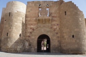 The image shows a historic stone gate with two large cylindrical towers on either side. The gate has an arched entrance with a metal gate, and above the arch, there is a decorative emblem or crest. The structure appears to be made of large stone blocks and has a weathered, ancient appearance. The sky is clear and blue in the background.