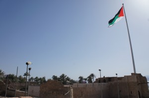 A large flagpole with the Palestinian flag is prominently displayed against a clear blue sky. Below the flag, there are stone buildings and structures, with some palm trees visible in the background. The scene appears to be in a historical or cultural site.