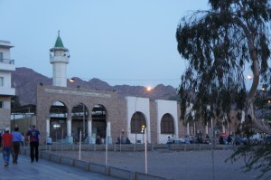 The image shows a mosque with a tall minaret topped with a green dome. The mosque has arched windows and doors, and it is situated in an urban area with a few people walking nearby. In the background, there are mountains and the sky appears to be at dusk or dawn. A tree is visible on the right side of the image.