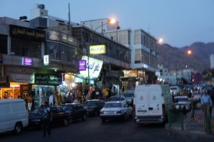 The image shows a busy street scene in the evening with several shops and buildings lining the street. There are multiple illuminated signs in various languages, and people are walking along the sidewalks. Several cars and vans are parked or driving on the street. The background features a mountainous landscape under a dusky sky. The overall atmosphere is lively and bustling.