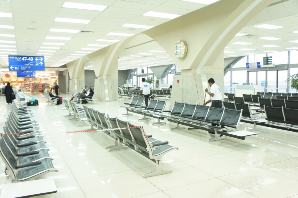 Alt text: The image shows a spacious airport waiting area with rows of empty black seats. A few people are seated in the background, some with luggage. A janitor is mopping the floor near the center of the image. Overhead, there are large, arched columns and a clock mounted on one of them. In the background, there is a blue sign displaying gate numbers 47-49 and directions in both English and Arabic. A small shop or kiosk is visible in the far left corner. The area is well-lit with a high ceiling and large windows on the right side.