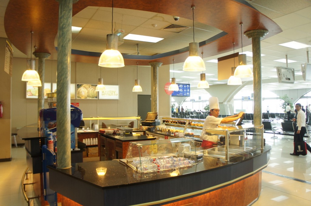 A bakery counter in an airport terminal. The counter is well-lit with hanging pendant lights and features a variety of baked goods on display. A staff member wearing a white chef's hat and uniform is working behind the counter. In the background, there are signs indicating gate numbers and directions, as well as seating areas for passengers. The overall atmosphere is clean and modern.