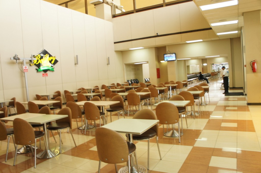The image shows an empty cafeteria with rows of tables and chairs arranged neatly. The chairs are brown and the tables have light-colored surfaces. There is a sign on the wall that reads "The Bakery" with a green banner underneath. The floor is tiled with a pattern of light and dark squares. In the background, there is a television mounted on the wall and a few people are visible, one standing and another sitting. The cafeteria has high ceilings and bright lighting.