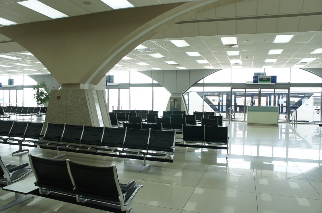 The image shows an empty airport waiting area with rows of black seats arranged in sections. The space is well-lit with large windows allowing natural light to enter. There are structural columns with an arch design, and a few digital screens displaying gate information. The floor is shiny and reflective, and there is a small counter or desk near the gates. The overall atmosphere is clean and modern.
