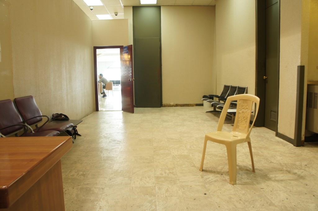 The image shows a waiting room with beige walls and a tiled floor. There are several chairs arranged along the walls. On the left side, there are maroon cushioned chairs, and on the right side, there are black plastic chairs. In the center of the room, there is a single beige plastic chair. At the far end of the room, there is an open door leading to another area where a person is seated at a table. The room is lit by overhead fluorescent lights.
