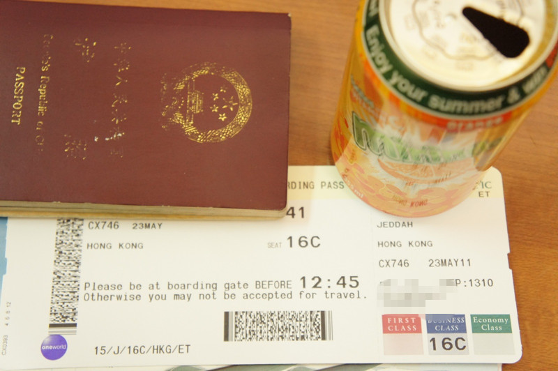 The image shows a Chinese passport, a boarding pass, and a can of drink on a wooden surface. The boarding pass indicates a flight from Jeddah to Hong Kong on May 23rd, with the flight number CX746 and seat 16C. The can of drink has colorful graphics and text on it.