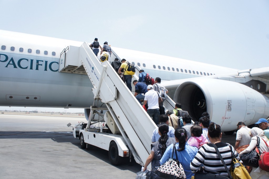 A group of people is boarding an airplane via a mobile staircase on an airport tarmac. The airplane has the word "PACIFIC" visible on its side, and the engine is marked with the Rolls-Royce logo. The sky is clear and the weather appears to be sunny. The passengers are carrying various bags and backpacks as they ascend the stairs.