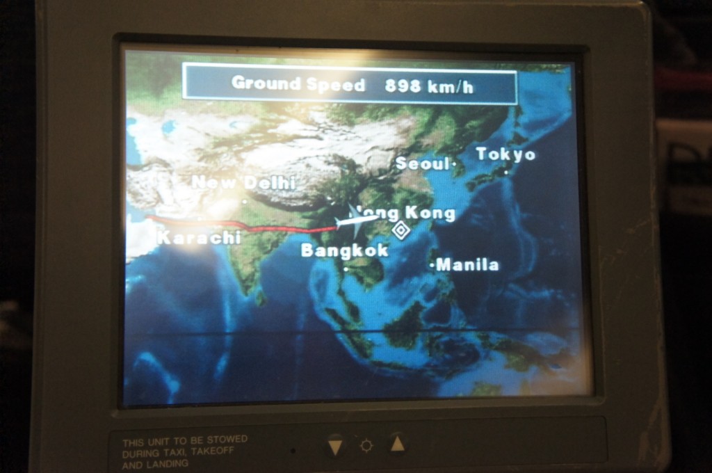 The image shows an in-flight entertainment screen displaying a flight path map. The map highlights several cities in Asia, including Karachi, New Delhi, Bangkok, Hong Kong, Seoul, Tokyo, and Manila. The flight path is marked with a red line, and the current ground speed of the aircraft is shown as 898 km/h. The screen is part of an airplane seatback entertainment system, with a note at the bottom indicating that the unit should be stowed during taxi, takeoff, and landing.