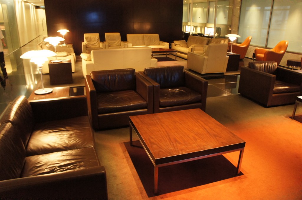 The image shows a modern, well-lit lounge area with various seating arrangements. There are several leather sofas and armchairs in dark brown and cream colors, arranged around wooden coffee tables. The space is illuminated by multiple table lamps with white shades, creating a warm and inviting atmosphere. The floor is a combination of carpet and glossy tiles, and the walls are dark, adding to the cozy ambiance. In the background, there are more seating areas and what appears to be a hallway leading to other parts of the building.