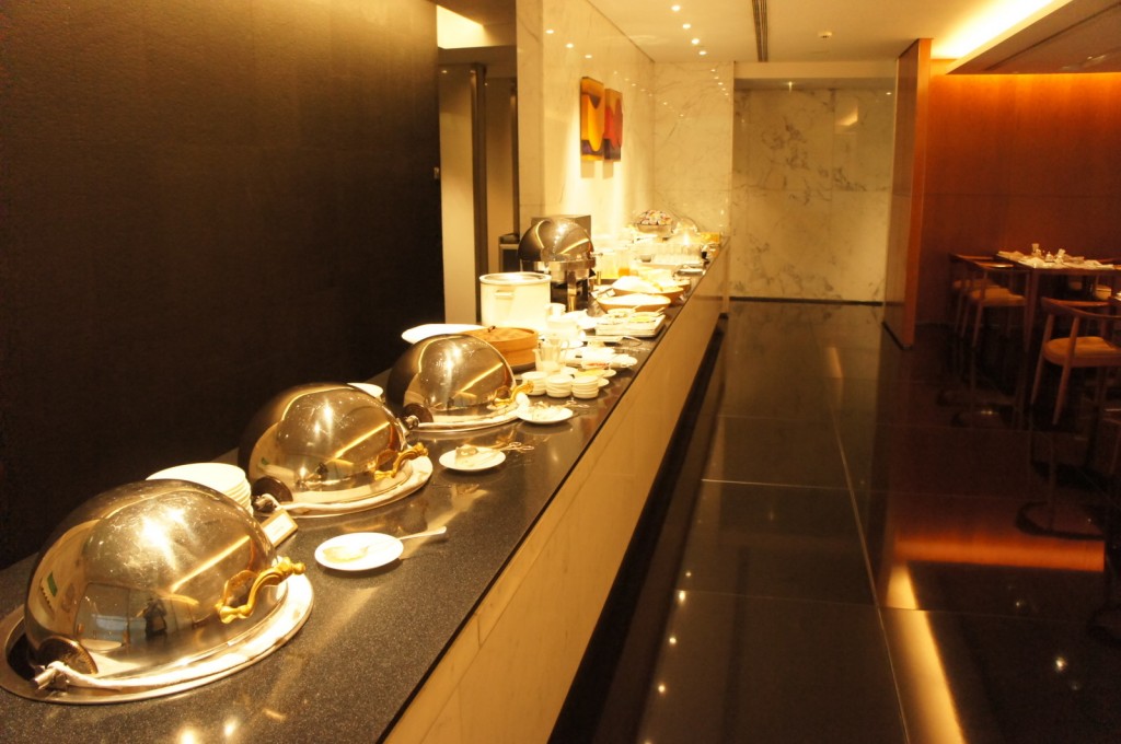 The image shows a buffet setup in a restaurant or hotel dining area. There are several covered chafing dishes lined up on a long counter, along with plates, bowls, and utensils. The counter is situated against a wall with a dark finish, and the floor is shiny and reflective. In the background, there are tables and chairs arranged for dining. The lighting is warm, creating a cozy atmosphere.