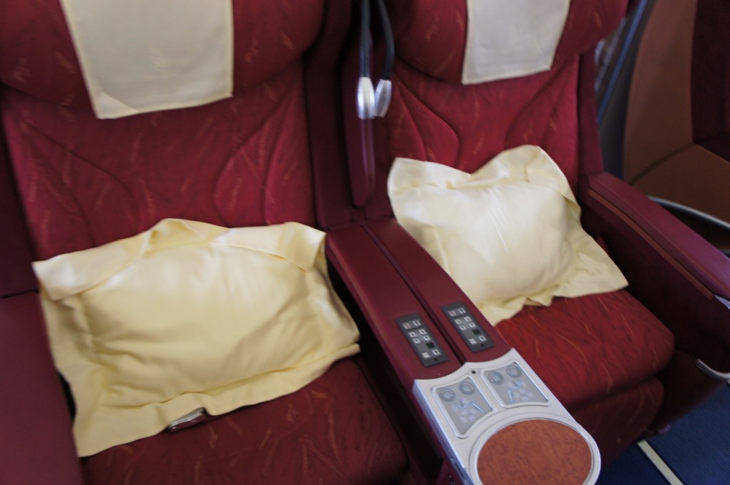 The image shows two adjacent red reclining seats, likely in a first-class or business-class section of an airplane. Each seat has a light yellow pillow and a headrest cover. Between the seats is a shared armrest with built-in control panels and a small wooden surface. The control panels appear to be for adjusting the seat positions and possibly other in-flight entertainment or comfort features.