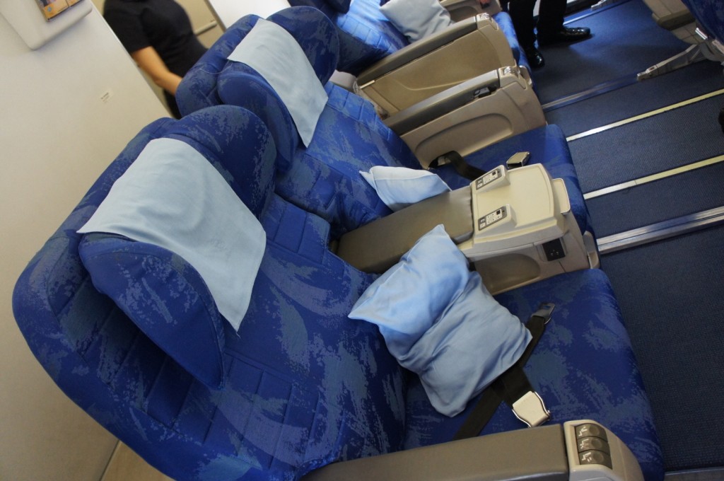 The image shows a row of blue airplane seats in a business or first-class cabin. The seats are wide and have light blue headrest covers and matching pillows. There are armrests with built-in controls and storage compartments between the seats. The cabin floor is carpeted in blue, and a person is partially visible in the background.