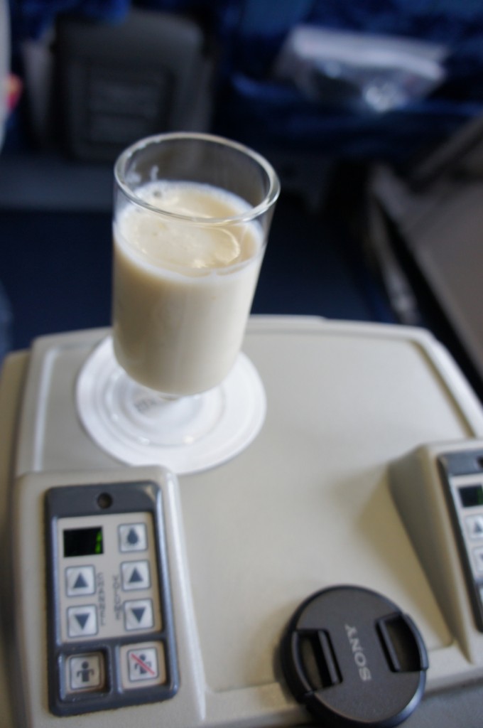 A glass of a creamy beverage, possibly a milkshake or smoothie, is placed on an airplane tray table. The tray table has control buttons on either side, and a camera lens cap labeled "Sony" is also visible on the table. The background shows airplane seats and part of the cabin interior.