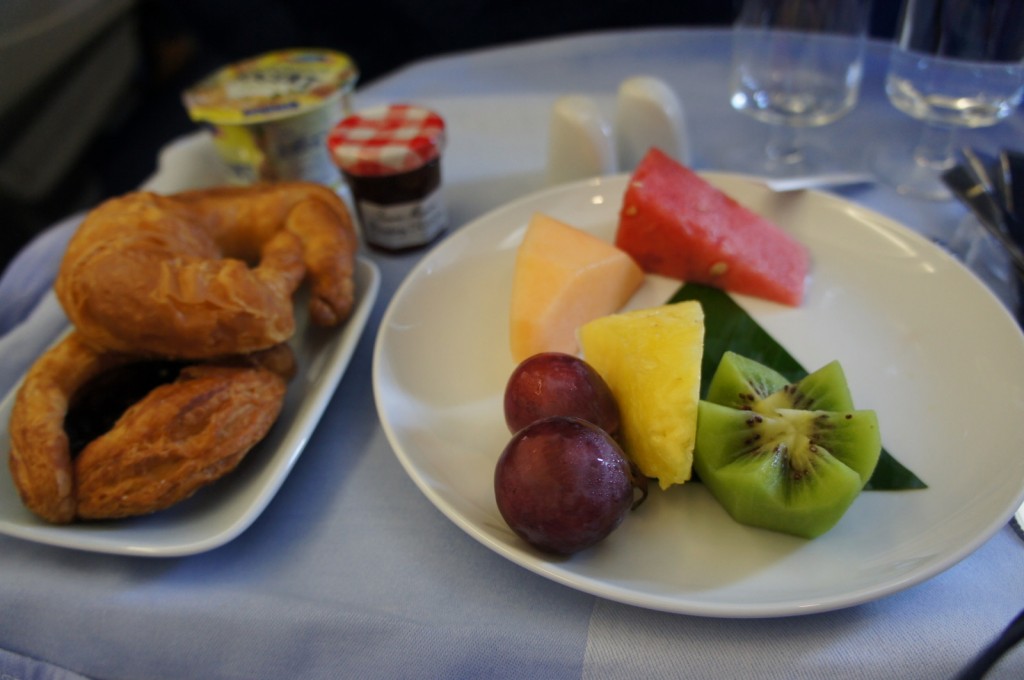 The image shows a meal consisting of a plate with various fresh fruits, including slices of watermelon, cantaloupe, pineapple, kiwi, and a few grapes. Next to it, there is a small plate with a croissant and a pastry. In the background, there is a small jar of jam and a container of yogurt. The setting appears to be on an airplane tray table, with a blue tablecloth and some glassware visible.