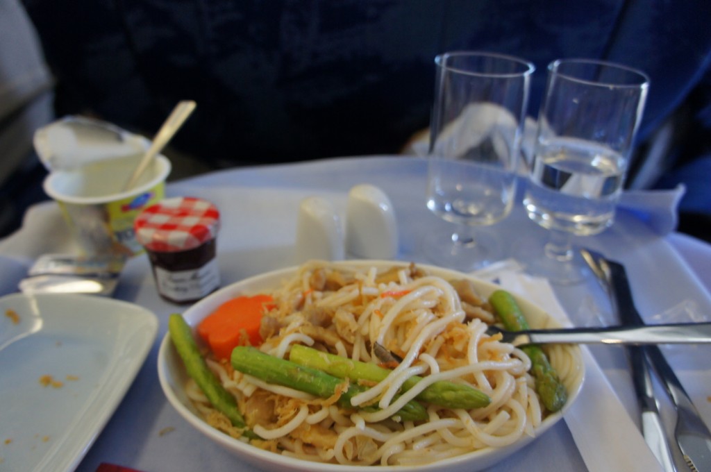 The image shows a meal served on an airplane tray table. The meal includes a bowl of noodles with vegetables such as asparagus and carrots. There are two glasses, one containing water, and the other empty. Additionally, there is a small container of yogurt with a spoon, a small jar of jam, and a set of cutlery including a knife and fork. The tray is covered with a white tablecloth.
