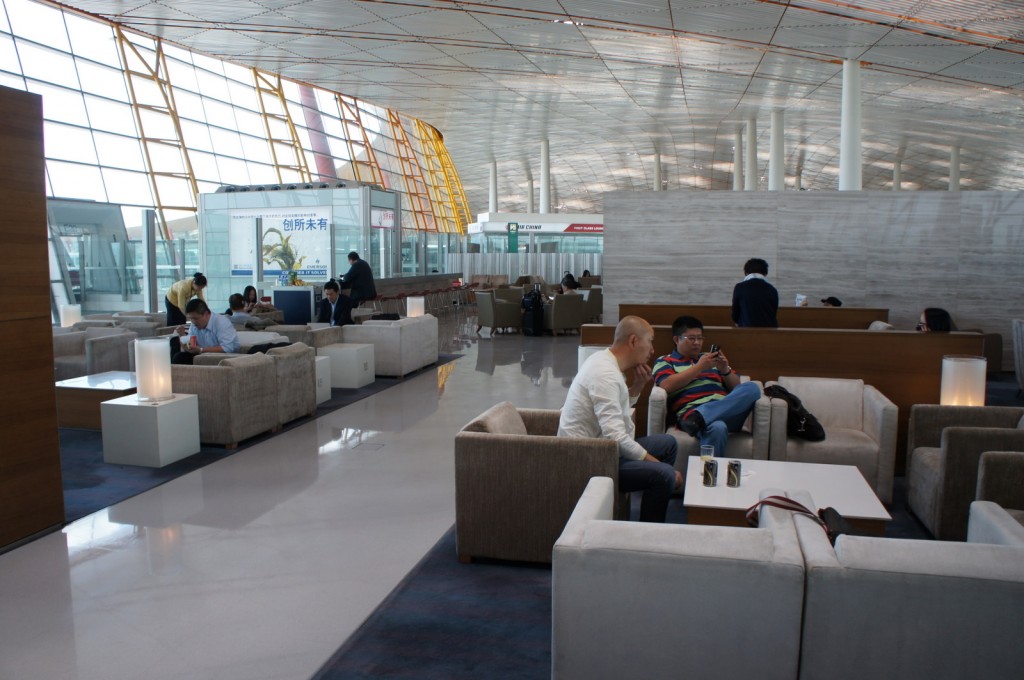 The image shows a modern airport lounge with several people seated on comfortable chairs and sofas. The lounge has a spacious and well-lit interior with large windows allowing natural light to enter. Some people are engaged in activities such as using their phones or laptops, while others are conversing. There are small tables with lamps placed between the seating areas. The ceiling has a sleek, curved design, and there are signs and advertisements visible in the background. The overall atmosphere appears to be calm and relaxed.