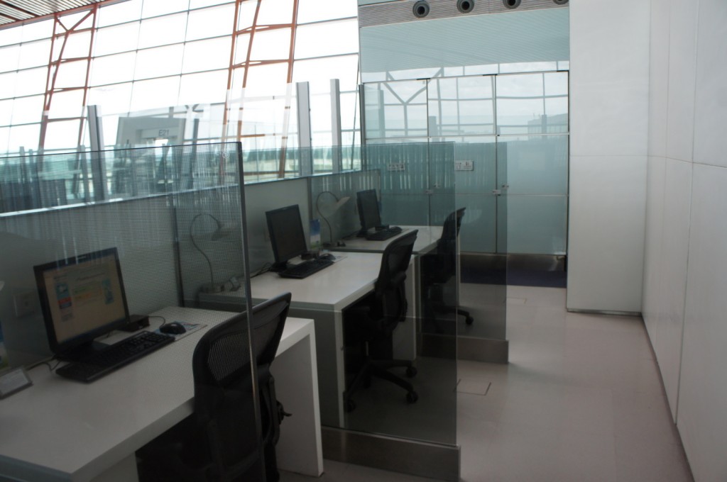 The image shows a modern office space with several workstations separated by glass partitions. Each workstation is equipped with a computer, a keyboard, a mouse, and an office chair. The area is well-lit with natural light coming through large windows in the background. The overall design is clean and minimalistic, with a focus on functionality and privacy for each workstation.