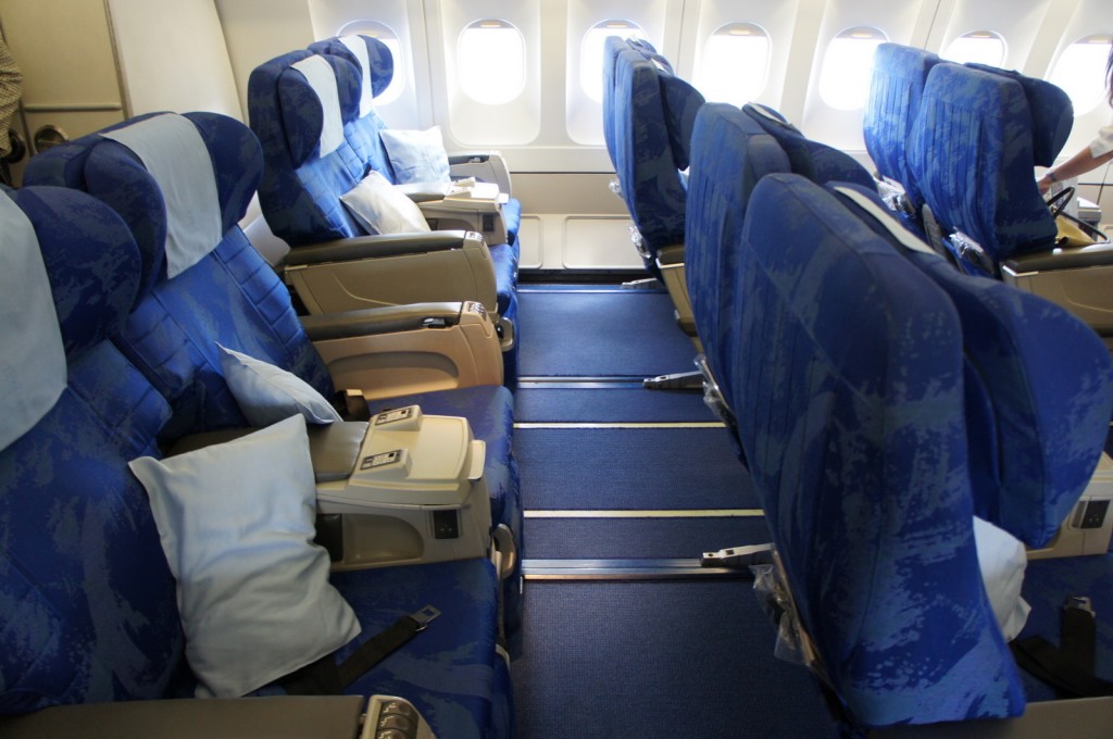 The image shows the interior of an airplane cabin, specifically the seating area. The seats are arranged in rows, with two seats on each side of the aisle. The seats are upholstered in blue fabric and have light blue headrest covers. Each seat has a pillow placed on it. The cabin windows are visible in the background, allowing natural light to enter. The armrests between the seats have built-in controls and tray tables. The overall setting appears clean and organized, ready for passengers.