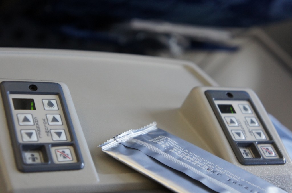 The image shows a close-up view of two control panels on the armrest of an airplane seat. Each panel has buttons for adjusting the channel and volume, as well as buttons for calling the flight attendant and a no-smoking indicator. There is also a packaged item, possibly a wet wipe or a similar amenity, placed on the armrest between the two control panels. The background is slightly blurred, indicating the interior of an airplane.
