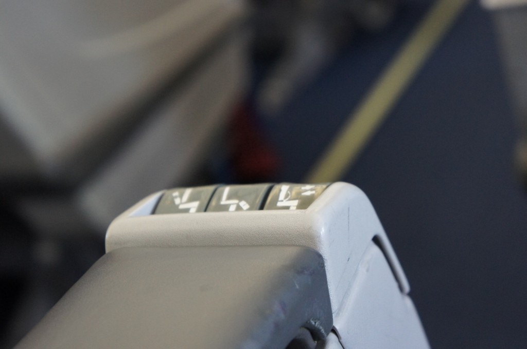 The image shows a close-up of an airplane seat armrest with control buttons. The buttons appear to be for adjusting the seat position, with symbols indicating different seat adjustments. The background is blurred, but it seems to be the interior of an airplane cabin.
