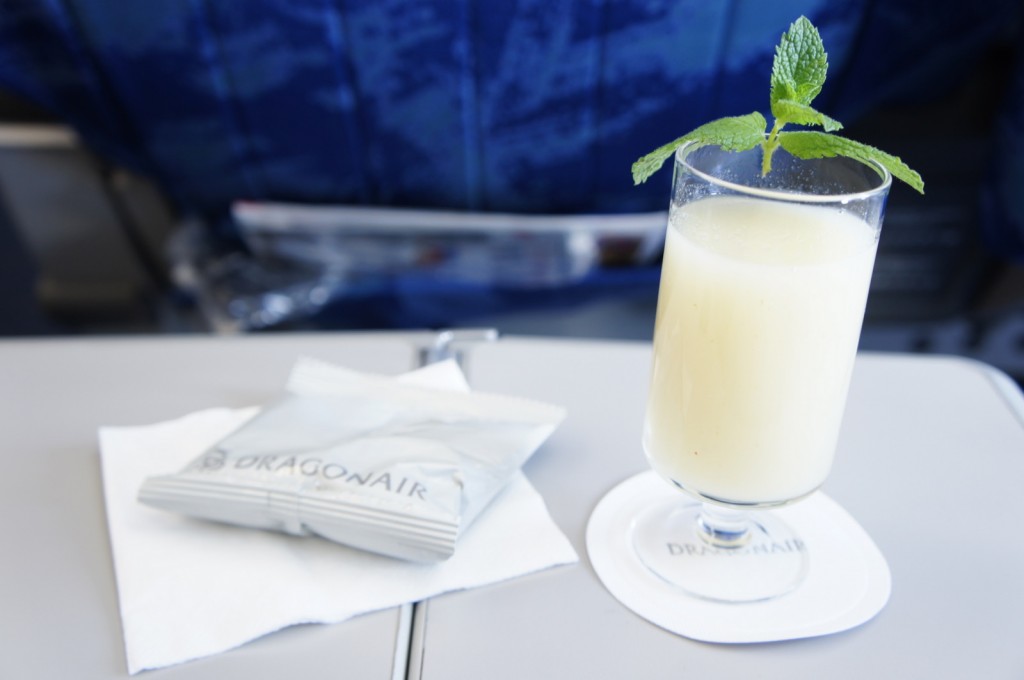 A glass of a creamy yellow beverage garnished with a sprig of mint is placed on a coaster on an airplane tray table. Next to the glass is a napkin and a packaged snack with the branding "Dragonair." The background shows the back of an airplane seat.