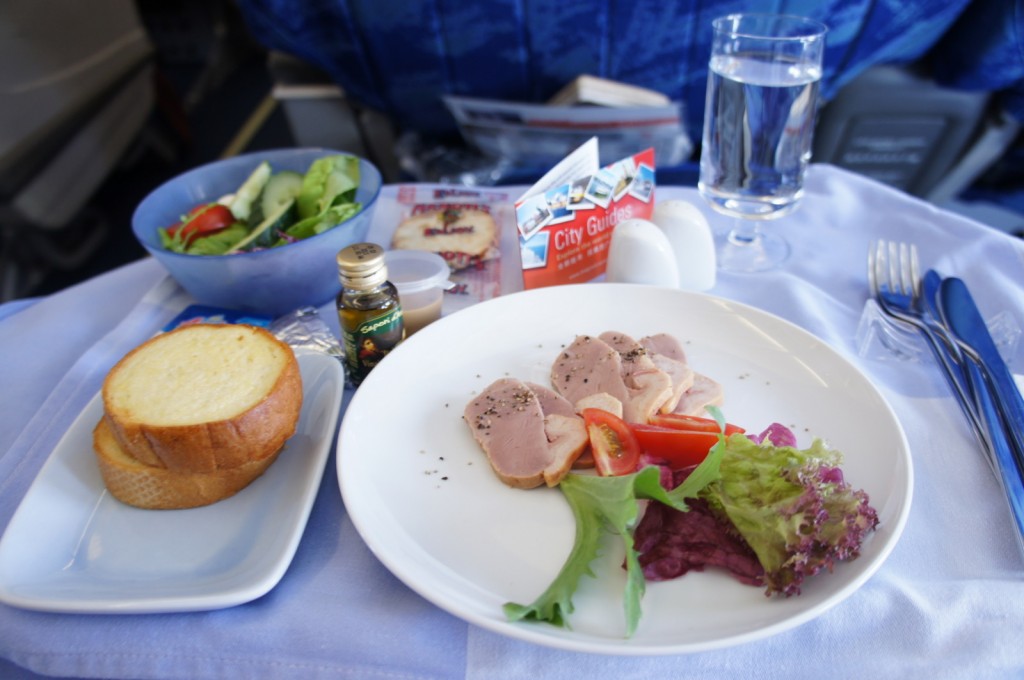 The image shows a meal served on an airplane tray table. The meal includes a plate with slices of meat, cherry tomatoes, and a small salad with lettuce. There is also a bowl of mixed salad with greens and tomatoes, a small bottle of olive oil, a bread roll, and a glass of water. Additionally, there are salt and pepper shakers, a set of cutlery wrapped in a napkin, and a city guide booklet on the tray. The background shows airplane seats and part of the cabin.