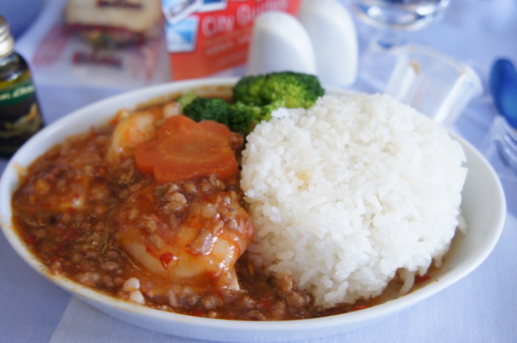 A bowl of food featuring a serving of white rice, a portion of minced meat in a red sauce, a slice of carrot, and some broccoli florets. The bowl is placed on a table with a blue tablecloth, and there are other items in the background, including a small bottle, a carton, and some salt and pepper shakers.