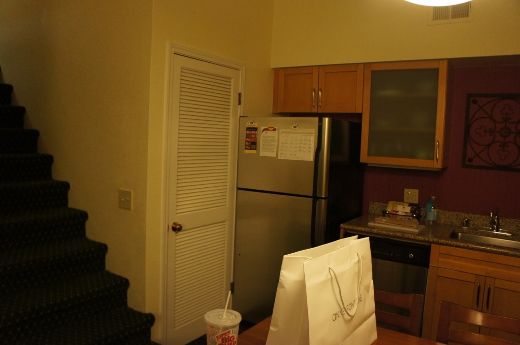 The image shows a small kitchen area with wooden cabinets, a stainless steel refrigerator, a sink, and a countertop. There is a white door with a louvered design next to the refrigerator. A staircase with dark carpeting is visible on the left side of the image. On the countertop, there are some items including a water bottle and a few other objects. In the foreground, there is a table with a white shopping bag and a large drink cup with a straw. The wall behind the sink has a decorative metal wall hanging.