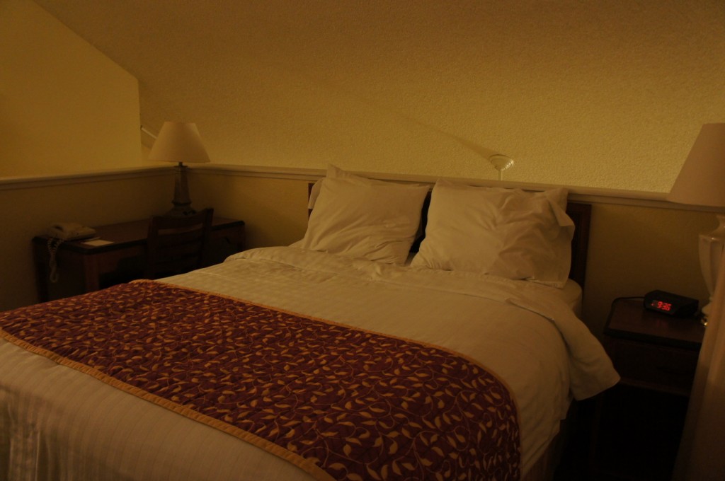 A cozy bedroom with a neatly made bed featuring white linens and a decorative maroon and gold patterned throw blanket. There are two pillows on the bed. On the right side of the bed, there is a nightstand with a lamp and an alarm clock displaying the time 9:36. On the left side, there is a small desk with a chair, a lamp, and a telephone. The room has a warm, dim lighting and a slanted ceiling.