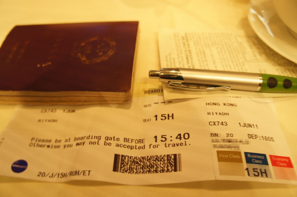 The image shows a close-up of a boarding pass, a pen, and a passport. The boarding pass indicates a flight from Hong Kong to Riyadh, with the flight number CX743 on June 1st. The seat number is 15H, and the boarding time is 15:40. The boarding pass also has a section indicating different classes: First Class, Business Class, and Economy Class, with the seat marked in Economy Class. The passport is partially visible and appears to be worn. The pen is silver with a green grip and is placed on top of some documents.