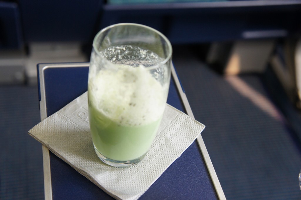 A glass of green-colored beverage with foam on top is placed on a white napkin on a tray table. The background appears to be the interior of an airplane.