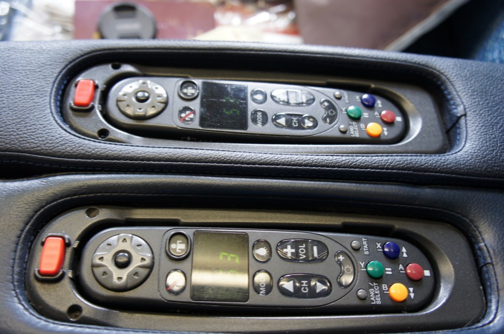The image shows two identical remote controls embedded in a leather surface, likely armrests. Each remote has a small screen, directional pad, and various buttons including volume, channel, and colored buttons (green, yellow, blue, and red). There is also a prominent red button on the left side of each remote. The remotes appear to be designed for controlling in-flight entertainment systems on an airplane.