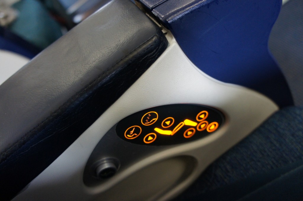 The image shows a close-up of an airplane seat armrest with a control panel. The control panel has several illuminated buttons with orange backlighting, which are used to adjust the seat position and other functions. The armrest is upholstered in dark leather or synthetic material.