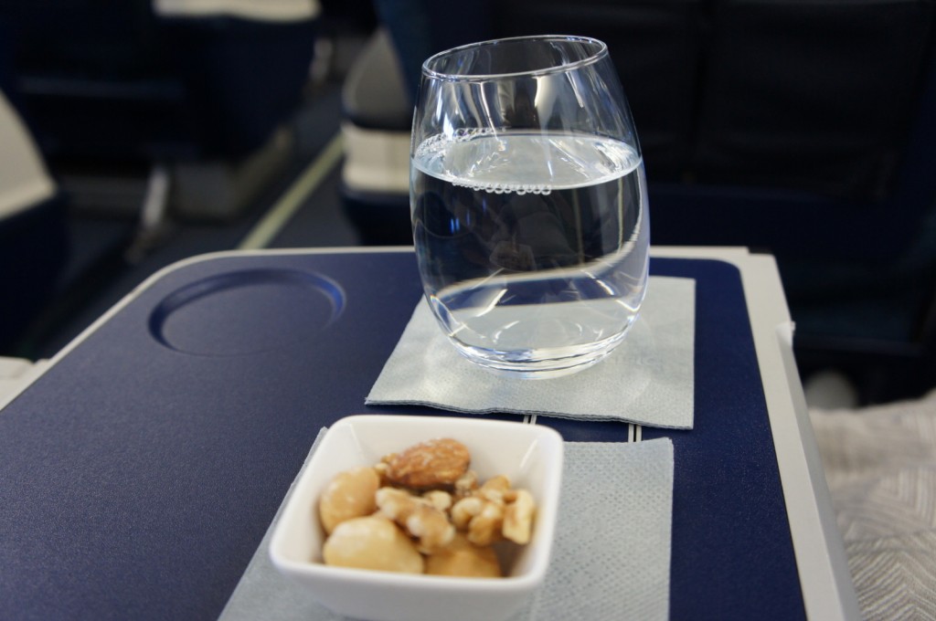 A glass of water and a small bowl of mixed nuts are placed on a tray table in an airplane. The glass and bowl are on separate napkins, and the background shows airplane seats.