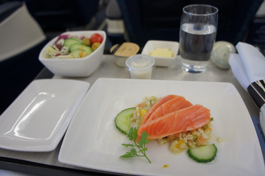 A meal served on an airplane tray table. The meal includes a plate with slices of salmon on a bed of grains and garnished with cucumber slices and a sprig of parsley. There is also a small bowl of salad with cherry tomatoes, cucumber slices, and other vegetables. Additionally, there is a glass of water, a small container of butter, a small cup of dressing or sauce, and a set of rolled-up napkins and utensils.