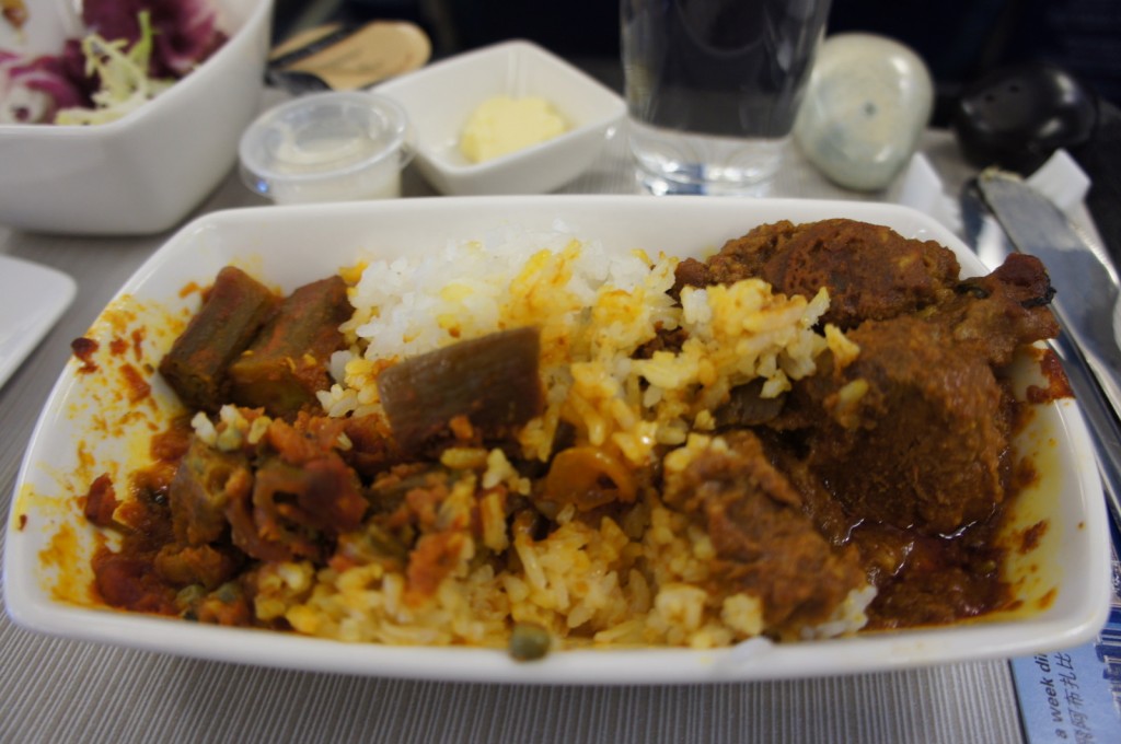 A rectangular white dish contains a meal of rice and a meat stew with pieces of okra. In the background, there is a small bowl of salad, a container of butter, a small cup of sauce, a glass of water, and some utensils. The setting appears to be a meal served on an airplane.