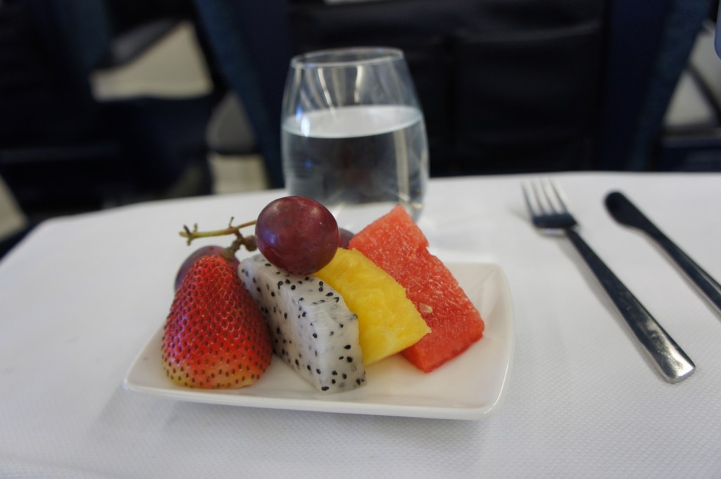 A small white plate with a variety of fresh fruits, including a strawberry, a piece of dragon fruit, a slice of pineapple, a piece of watermelon, and a red grape on top. The plate is placed on a white tablecloth, with a glass of water, a fork, and a knife in the background. The setting appears to be on an airplane tray table.