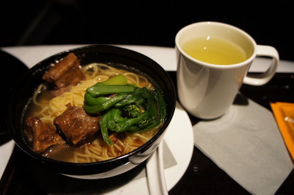 A bowl of noodle soup with chunks of meat and green vegetables, accompanied by a cup of tea. The bowl is placed on a white plate with a napkin underneath, and the cup of tea is on the right side of the bowl.