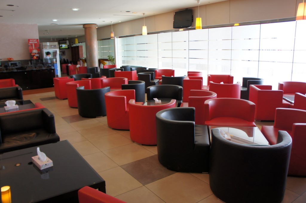 The image shows a modern, well-lit lounge area with numerous red and black armchairs arranged around small glass-topped tables. The space has a clean, contemporary design with large windows along one side, allowing natural light to fill the room. There are pendant lights hanging from the ceiling, and a television mounted on the wall. The lounge appears to be a comfortable and inviting place for people to sit and relax.