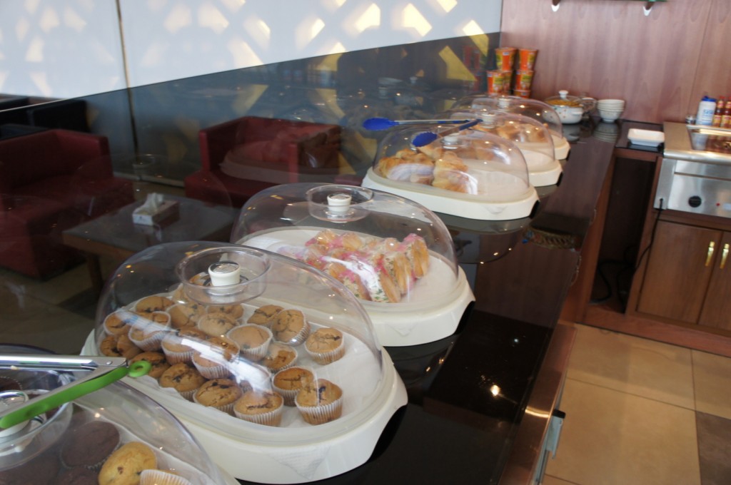 The image shows a buffet setup with various baked goods displayed under clear plastic domes. The items include muffins, pastries, and sandwiches. The buffet is set on a countertop in a room with red chairs and a wooden cabinet in the background. There are also some bowls and canned goods on the counter.
