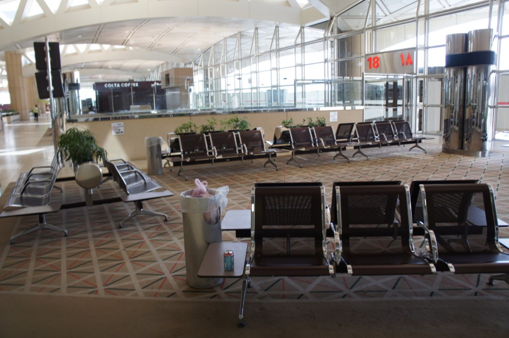The image shows an airport waiting area with rows of empty seats. In the background, there is a Costa Coffee shop. The area is well-lit with natural light coming through large windows. There are some plants placed around the seating area, and a trash can with a plastic bag and a can of soda on a small table in the foreground. The gate numbers 18 and 1A are visible on the right side of the image. The floor has a patterned carpet.