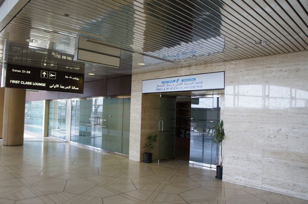 The image shows the entrance to a "First & Business Class Lounge" at an airport. The entrance is marked by a sign above the door with text in both English and Arabic. The door is made of glass, and there is a potted plant to the right of the entrance. Above the entrance, there is a ceiling with a metallic finish. To the left, there is a directional sign indicating gates 21-28 and the "First Class Lounge" in both English and Arabic. The floor has a geometric tile pattern.