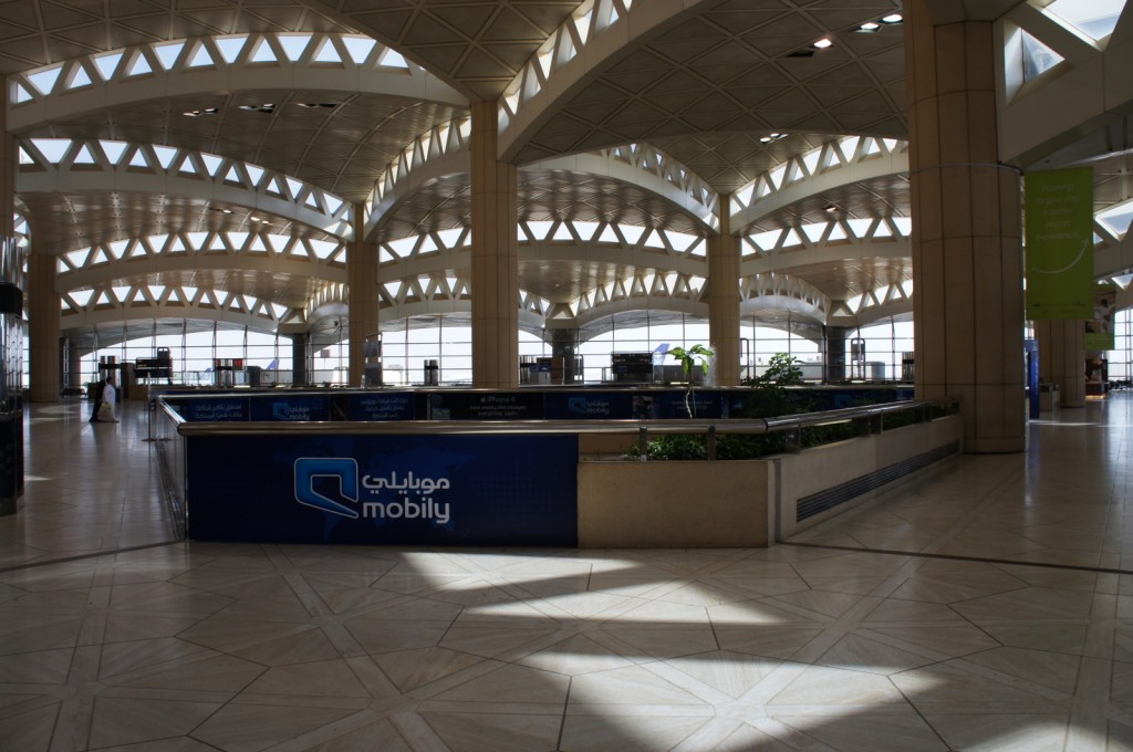 The image shows the interior of an airport terminal with a modern architectural design. The ceiling features a series of triangular skylights that allow natural light to illuminate the space. There are large columns supporting the structure and a spacious, open floor plan. In the center of the image, there is a blue advertisement banner with the logo and text for "Mobily," a telecommunications company. The terminal appears to be relatively empty, with only a few people visible in the background. There are also some green plants in planters, adding a touch of greenery to the space.