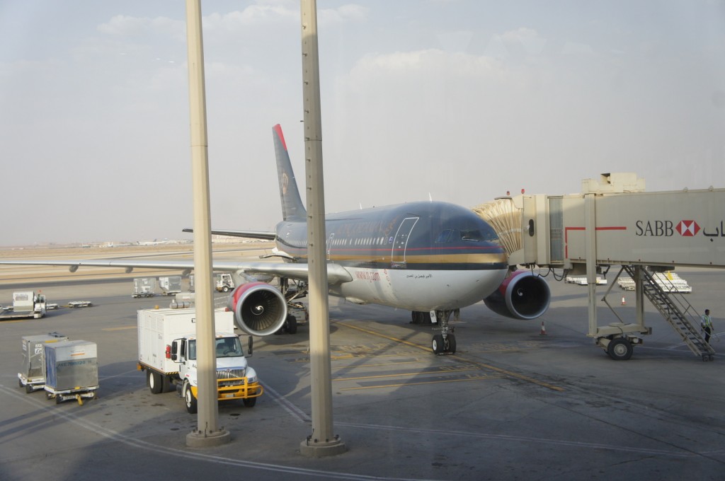 The image shows a commercial airplane parked at an airport gate. The aircraft is connected to a jet bridge on the right side, which is branded with the logo and name "SABB." There are various ground service vehicles and equipment around the plane, including a white truck and luggage carts. The scene appears to be taken from inside the airport terminal, looking out through large windows. The sky is partly cloudy, and the overall atmosphere suggests a typical day at the airport.