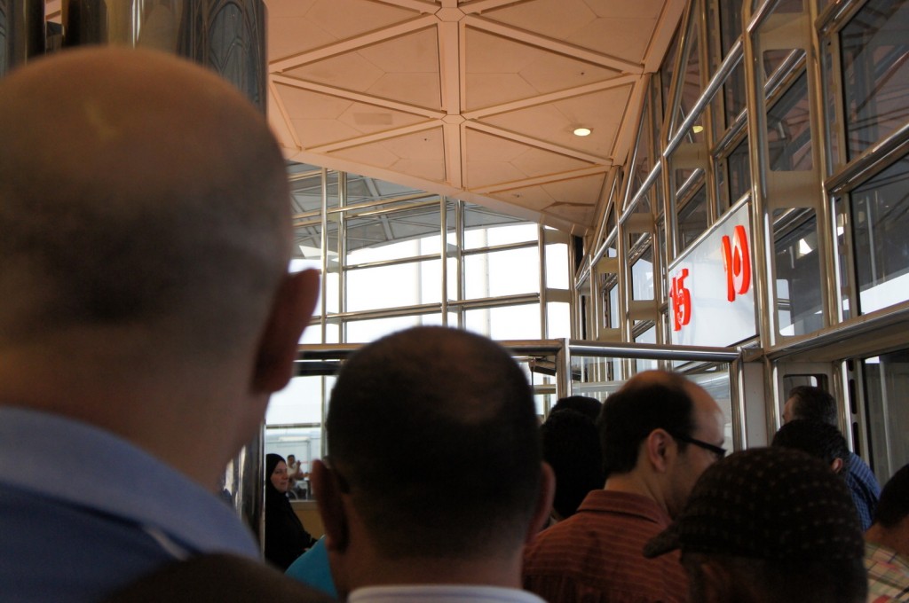 A group of people is standing in line inside a building with glass walls and a geometric ceiling. The focus is on the backs of several individuals, some of whom are bald or have short hair. In the background, there is a sign with the numbers "15" and "10" in red. The atmosphere suggests a public place, possibly an airport or a train station.