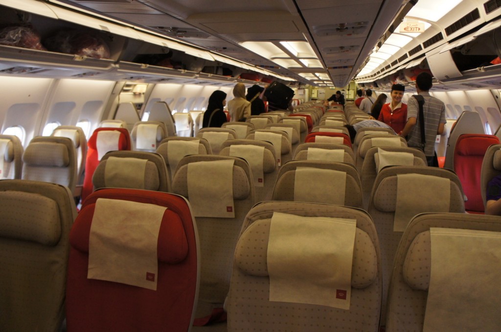 The image shows the interior of an airplane cabin. The seats are arranged in rows, with some seats having red covers and others having beige covers. Each seat has a headrest cover. Overhead compartments are visible, some of which are open with luggage inside. Several passengers are standing in the aisle, some placing their luggage in the overhead bins. The lighting is bright, and the cabin appears to be preparing for takeoff or landing.