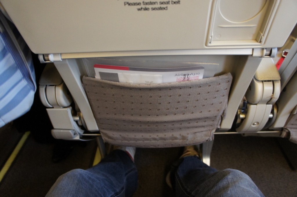 The image shows the view from a passenger's seat on an airplane. The back of the seat in front is visible, including a seat pocket containing a safety instruction card for an Airbus A310. The passenger's legs and feet are also visible, with the person wearing jeans and sneakers. The text "Please fasten seat belt while seated" is printed on the back of the seat.