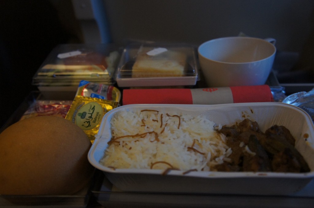 The image shows an airline meal tray. The tray contains a main dish with white rice and a meat or vegetable curry, a bread roll, a small container of butter, a dessert in a plastic container, a cup, and a red and white napkin. There are also some additional items in the background, possibly condiments or utensils.