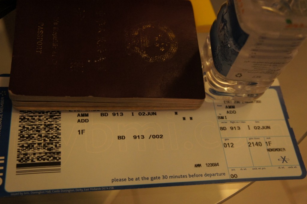 The image shows a close-up of a traveler's items on a surface. There is a passport, a boarding pass, and a bottle of water. The boarding pass indicates a flight from AMM (Amman) to ADD (Addis Ababa) on flight BD 913, with a departure date of June 2nd. The seat number is 1F, and the gate is 012 with a departure time of 21:40. The passport is partially covering the boarding pass, and the water bottle is placed to the right of the boarding pass.